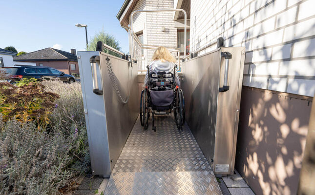 Frau im Rollstuhl fährt auf Hublift im Außenbereich eines Einfamilienhauses.